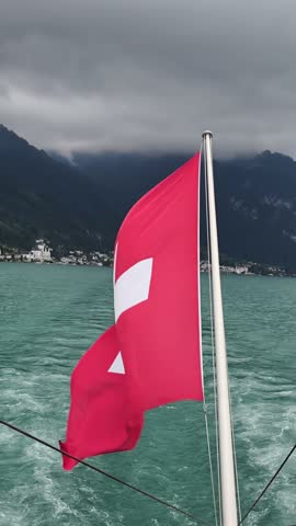 Swiss national flag waving at the stern of a boat cruising across the turquoise waters of Lake Lucerne. In the background, the dramatic Swiss Alps rise into low clouds, creating a moody and atmospheri
