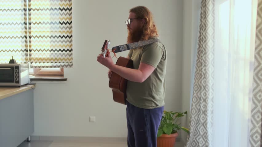 A modern indie artist plays acoustic guitar in his kitchen, focused on crafting a new track for social media. Stylish home vibes