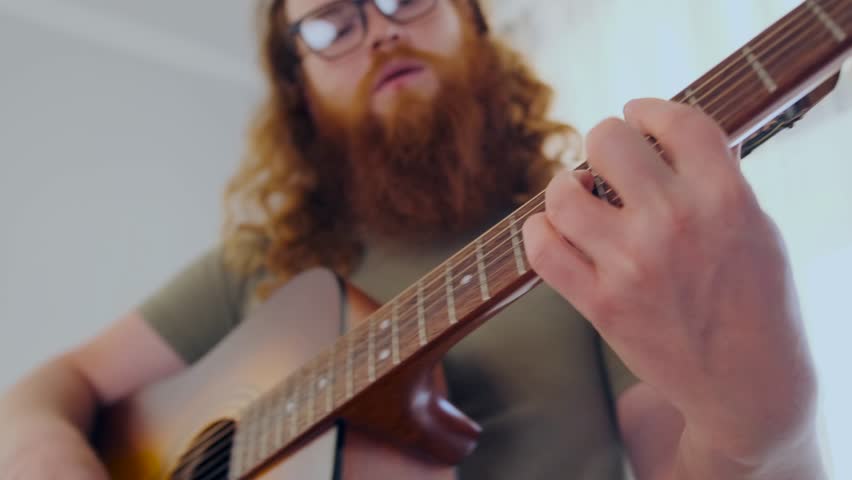A soulful male bard with long red curls plays a heartfelt tune on acoustic guitar in his cozy room, searching for lyrics and harmony in silence.