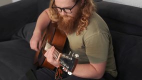 A man with curly hair and beard sits on a couch in a home studio, gently strumming his guitar and swaying to the rhythm. Composes an original melody, getting ready to record a video for social media. - Powered by Shutterstock - Get 15% off with code: PIKWIZARD15