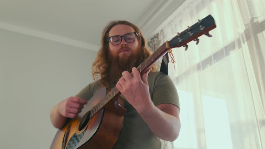 Low-angle shot of a bearded man with curly red hair and glasses rhythmically playing an acoustic guitar while standing near a bright window. Indie music and creative lifestyle themes.