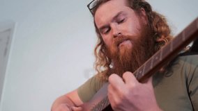 Close-up portrait of a young man with long curly red hair, glasses, and a thick beard, deeply focused while playing acoustic guitar at home. Musical inspiration, creativity and emotional expression. - Powered by Shutterstock - Get 15% off with code: PIKWIZARD15