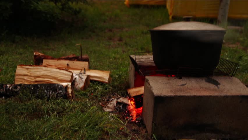 A cozy scene of camping in nature, with a cauldron of food cooking over a roaring fire. The nearby wood and firewood create a warm and inviting atmosphere, perfect for a peaceful travel adventure.