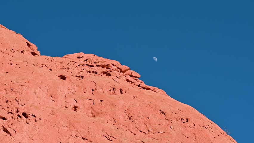 Aerial shot of moon rising over sunlit red rock ridge in Garden of the Gods, Colorado, with clear blue sky backdrop.