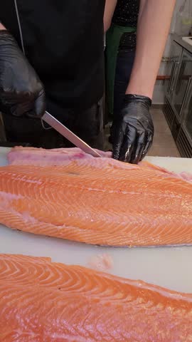 Chef slicing fresh salmon fillet with precision on a cutting board in a professional kitchen. Close-up of hands in black gloves and raw fish texture
