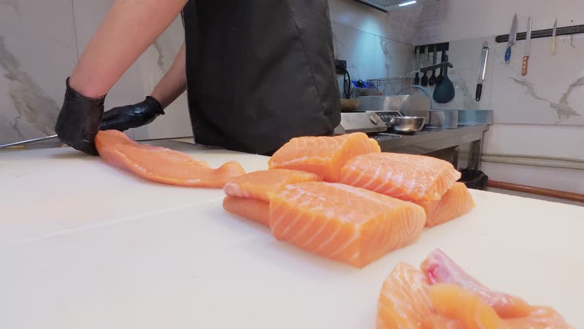 Chef slicing fresh salmon fillet with precision on a cutting board in a professional kitchen. Close-up of hands in black gloves and raw fish texture