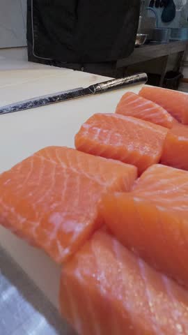 Chef slicing fresh salmon fillet with precision on a cutting board in a professional kitchen. Close-up of hands in black gloves and raw fish texture