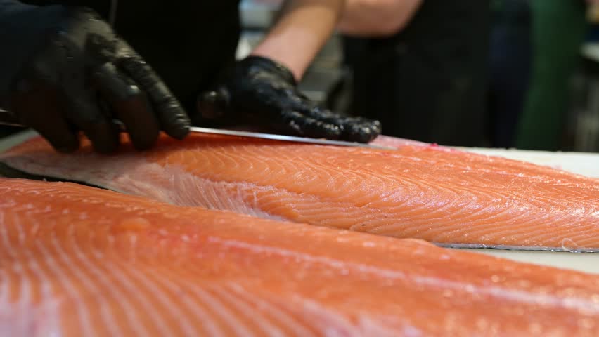 Chef slicing fresh salmon fillet with precision on a cutting board in a professional kitchen. Close-up of hands in black gloves and raw fish texture