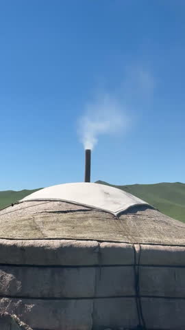 A traditional yurt with a smoke plume rising from its chimney under a bright blue sky, showcasing cultural architecture against a natural backdrop of hills, Mongolia, tc01