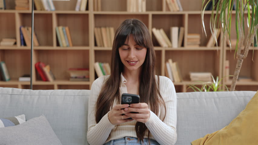 Gorgeous Caucasian woman sitting on sofa and smiling warmly while holding smartphone with both hands. Looking directly at camera. Waiting for response while staying relaxed in book-filled living room.