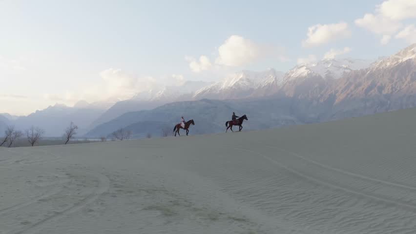 Aerial View of Horseback Riders on Sand Dunes in Pakistan, with Majestic Snow-Capped Mountains in the Background
