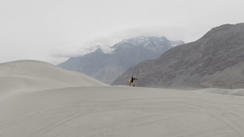 A lone rider traverses the vast sand dunes of Pakistan, with majestic snow-capped mountains