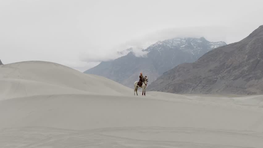 A lone rider traverses the vast sand dunes of Pakistan, with majestic snow-capped mountains in the distance