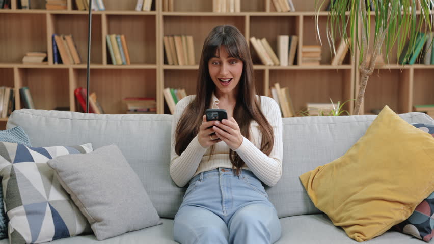 Excited Caucasian woman celebrating while holding smartphone, raising fist in joy and sticking tongue out playfully. Sitting on couch in light-filled room with book-filled shelves in background.