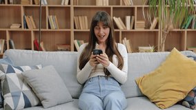 Excited Caucasian woman celebrating while holding smartphone, raising fist in joy and sticking tongue out playfully. Sitting on couch in light-filled room with book-filled shelves in background. - Powered by Shutterstock - Get 15% off with code: PIKWIZARD15