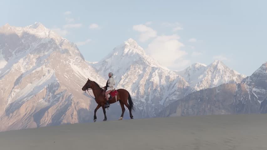 A lone rider traverses the vast sand dunes of Pakistan, with majestic snow-capped mountains forming a breathtaking backdrop
