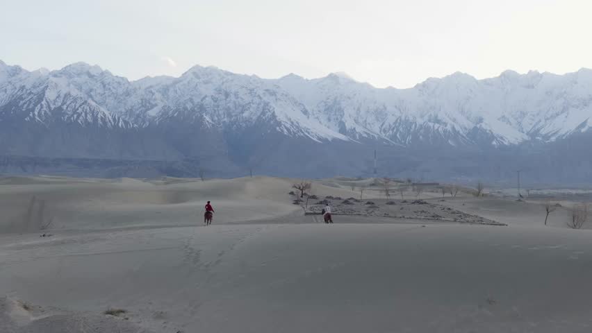 Aerial view of a desert landscape in Pakistan, featuring snow-capped mountains and horseback riders