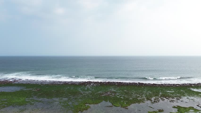 Vertical 9:16 drone footage, 2K 30fps. Aerial follow shot of green waves crashing over coral rocks along the shoreline. Dynamic coastal view with natural textures and ocean movement.