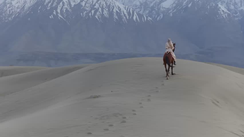 A lone rider traverses the vast sand dunes of Pakistan, with majestic snow-capped mountains in the backdrop