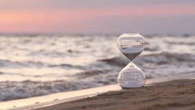 White sand hourglass resting on sandy beach, gentle sea waves rolling in background during soft, pale sunset, symbolizing time passing and tranquil moments of reflection. Selective focus shot - Powered by Shutterstock - Get 15% off with code: PIKWIZARD15