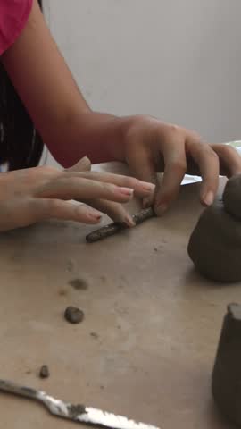 Hands delicately shaping a thin cylindrical clay form on a table, with a rounded clay base nearby. The close-up shows precision work in forming components for a larger artistic piece.