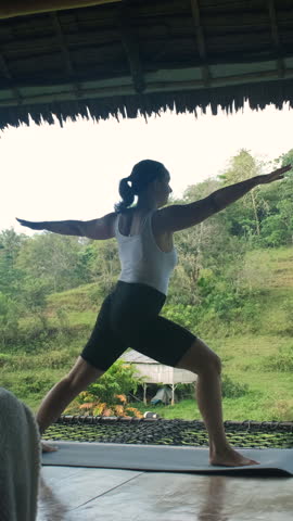 Vertical video. Woman gracefully performs yoga on bamboo hut terrace with vibrant greenery and rice fields, slow motion 