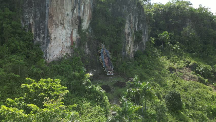 Bright sunny day aerial of the Nuestra Señora de Guadalupe statue on rocky terrain in Río San Juan, Dominican Republic, framed by rich green vegetation. Suited for cultural, nature, and tourism videos
