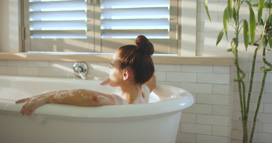 Woman leaning back, tilting head lifting leg in freestanding tub for calm bubble bath, copy space. Relaxation, wellness, serenity, minimalist, tranquil, selfcare, interior