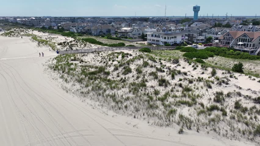 Aerial view of Stone Harbor, NJ. Elegant beachfront homes and coast behind wide sandy beach with dunes, umbrellas and walkways. Classic East Coast summer scene under clear skies. Wide shot.