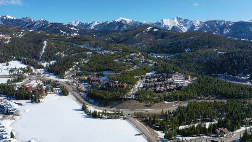 Aerial footage shows winter condos in Big Sky, Montana, dusted in snow and nestled among tall pines, with quiet streets and panoramic mountain backdrops in a calm, scenic town setting.