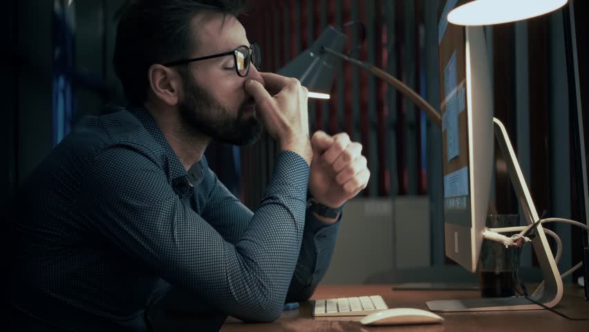 Man With Eye Strain At Computer. Tired Professional Working Late. Overworked Employee Digital Fatigue. Long Hours At Desk. Eye Rubbing Gesture. Mental Health. Stressful Work Environment Exhaustion
