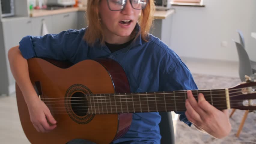 Close-up of a young woman singing emotionally while playing acoustic guitar in her kitchen. Intimate atmosphere, expressive face, and fingers on strings in focus.