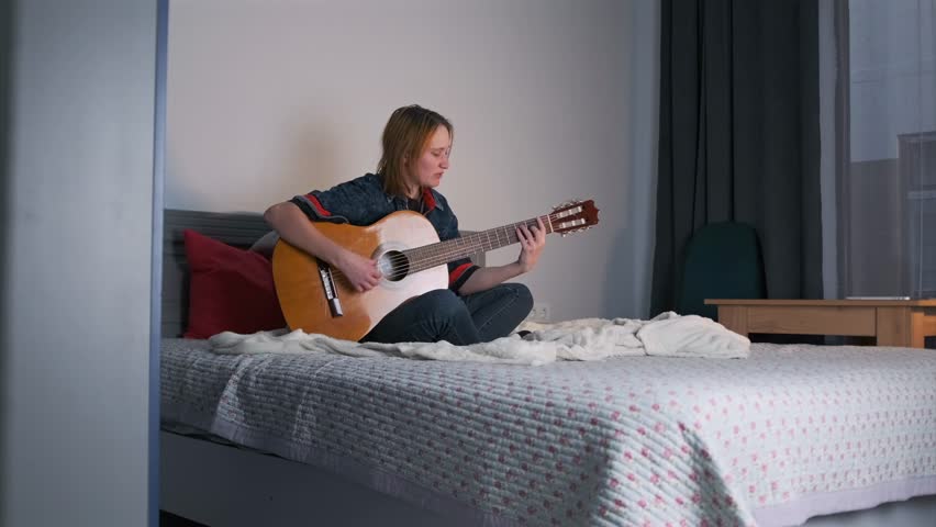 Wide shot of a young woman sitting cross-legged on a bed, singing while playing an acoustic guitar. Casual outfit, soft daylight, and cozy bedroom setting, home music moment.