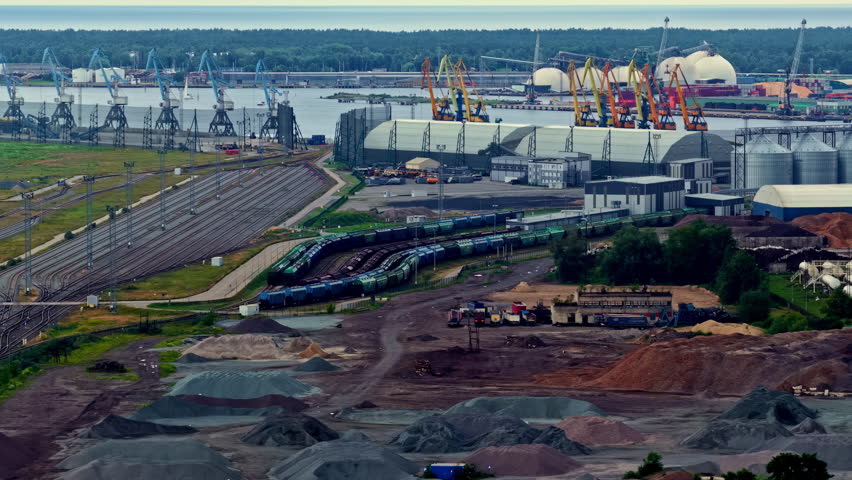 An Industrial Port Complex Stretches Along the Waterfront, with Cargo Trains, Cranes, Silos, and Stockpiles of Raw Materials Spread Across the Site - Aerial Drone Shot