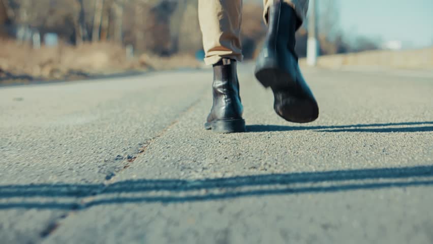 Feet In Black Boots Walking On Asphalt Road. Person Strolling Outdoors. Urban Exploration, Adventure And Travel. Pavement Walk Modern Footwear. Walking Away Independent Spirit. Walking Alone Footwear