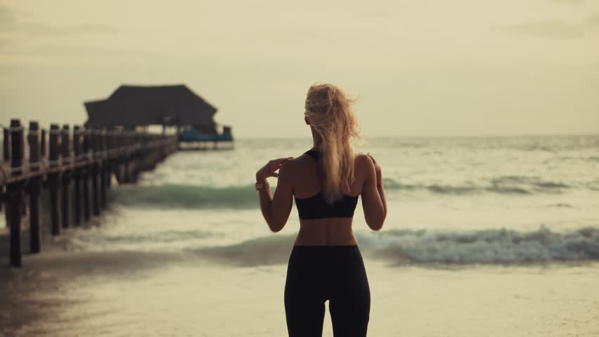 Blonde Woman Stretching Arms At Beach. Female Exercising Outdoors. Sunset Workout Fitness And Wellness. Beach Exercise Vacation Fitness. Healthy Living Warm-Up Routine. Morning Stretch Exercise Habits