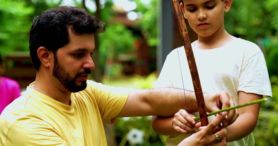 Indian father teaching archery to son using homemade bow and arrow in home garden, spending quality family time together playing outdoor sports, learning skills, bonding through fun activity