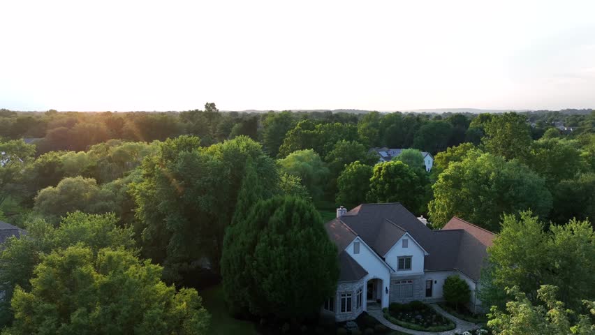 Classic American suburban house with pitched roof, gables and large windows. Aerial view at sunrise highlights traditional architecture surrounded by lush trees. Aerial top down. Quiet atmosphere.