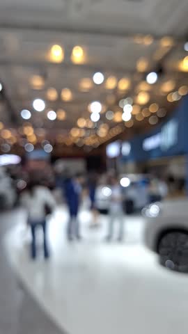 Blurry view of a large crowd walking at the Auto Show inside the convention hall, with vibrant lights and displays in the background, creating a lively and busy bokeh effect.