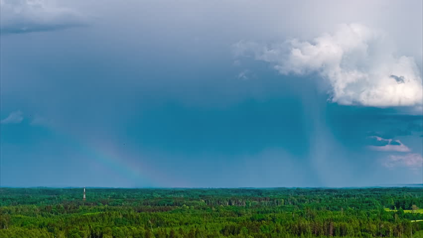 A beautiful aerial timelapse captures a faint rainbow appearing under dramatic rain clouds as they move across the sky above a vast and dense green forest.