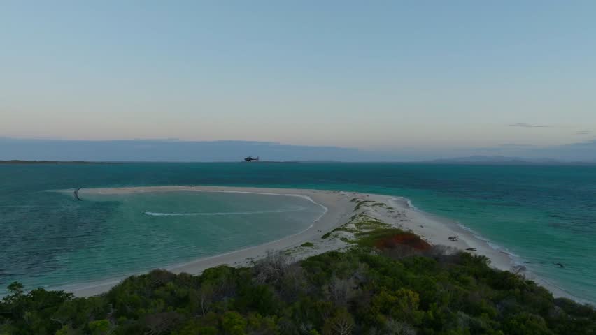 Fast drone orbit reveals Private island in northeast Madagascar, showcasing white sandbars, turquoise waters, and scattered vegetation in the soft evening light under a pale blue sky.