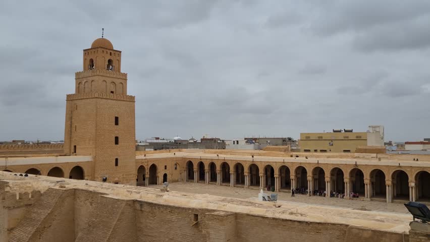 Panoramic view of the minaret, walls, and courtyard of the famous Great Mosque of Kairouan on a partly cloudy day in Tunisia, showcasing its historic Islamic architecture