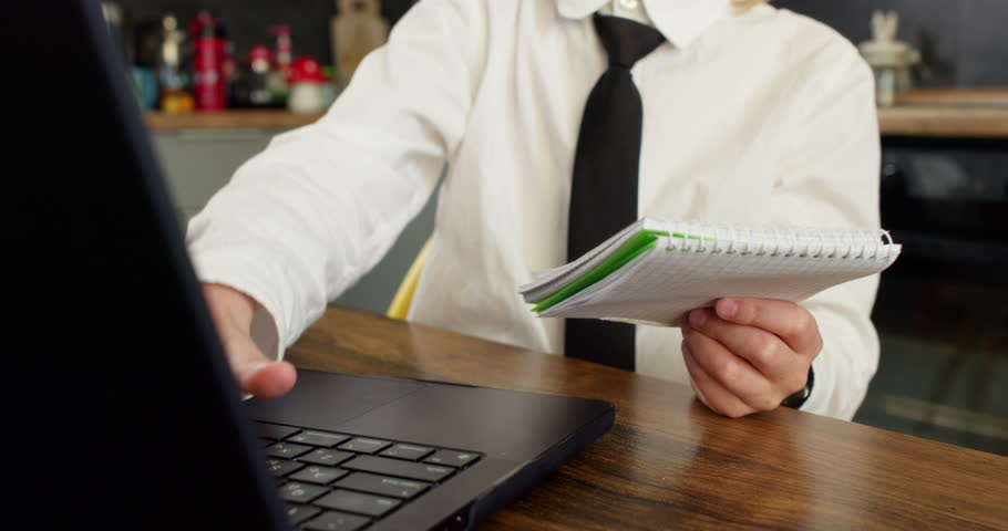 Boy in white shirt and black tie types on laptop keyboard while holding small spiral-bound notebook. Focus on engagement with digital and physical tools for study
