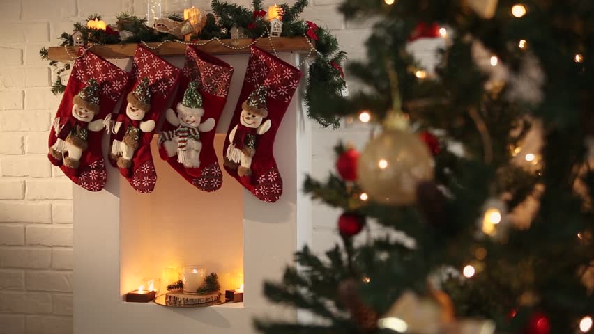 A cozy Christmas scene featuring a decorated fireplace with red stockings hanging. A Christmas tree adorned with lights and ornaments is visible in the background.