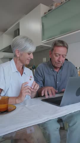 Senior couple navigating online shopping, experiencing digital learning challenges while using laptop and credit card in home kitchen setting
