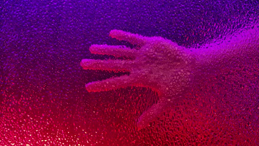 Close up of a hand pressed against frosted glass, illuminated by vivid red and purple lighting. The textured surface distorts details, creating an artistic, moody effect.