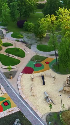 A stunning vertical aerial parallax shot reveals a vibrant park with modern playgrounds in Latvia, surrounded by green trees and a peaceful cityscape in summer sunlight.