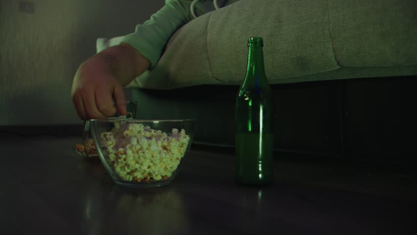Close up of a male hand greedily reaching for popcorn in a glass bowl standing on the floor next to a sofa and a green bottle in a living room at night. Lazy lifestyle and bad habits of adult man