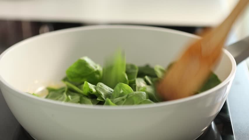 Close-up of female hands frying fresh spinach in a pan at home kitchen. Healthy vegetarian meal preparation with organic greens in a rustic cooking setting.