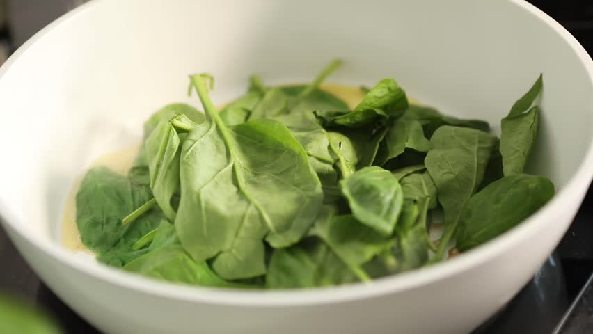 Close-up of female hands frying fresh spinach in a pan at home kitchen. Healthy vegetarian meal preparation with organic greens in a rustic cooking setting.